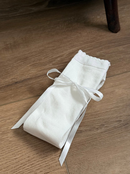 A pair of Historically Inspired Cotton Knee-High Socks with Satin Ribbon Lacing in Off-White, pictured on a historic wooden cottage floor.