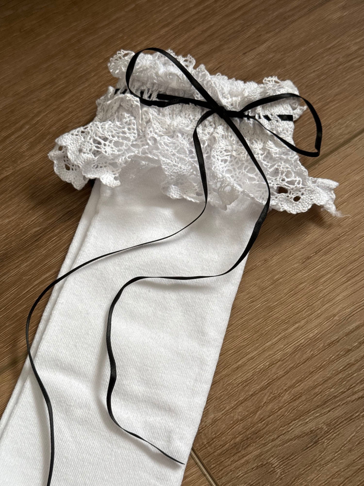 Historically Inspired white Cotton Knee-High Socks with ruffled Lace Trim and black satin ribbon ties on a cottagecore wooden floor.