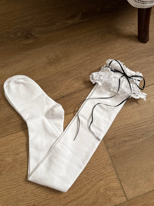 Historically Inspired white Cotton Knee-High Socks with ruffled Lace Trim and black satin ribbon ties on a cottagecore wooden floor.