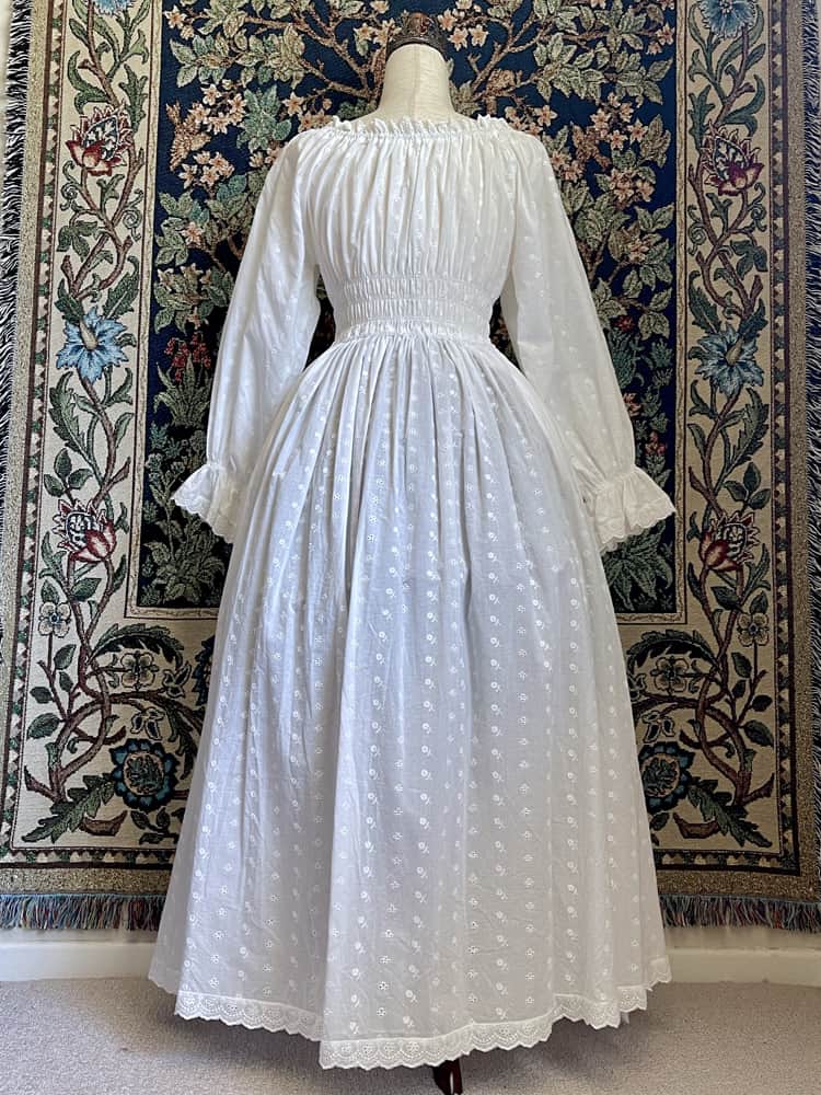 A Historically inspired white 100% cotton smocked chemise dress with bishop bell sleeves is pictured on a mannequin in front of an ornate tapestry background.
