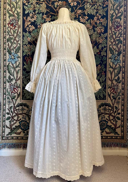 A Historically inspired sheer ivory 100% Cotton smocked chemise maxi dress with bishop bell sleeves is pictured on a mannequin in front of an ornate botanical tapestry.