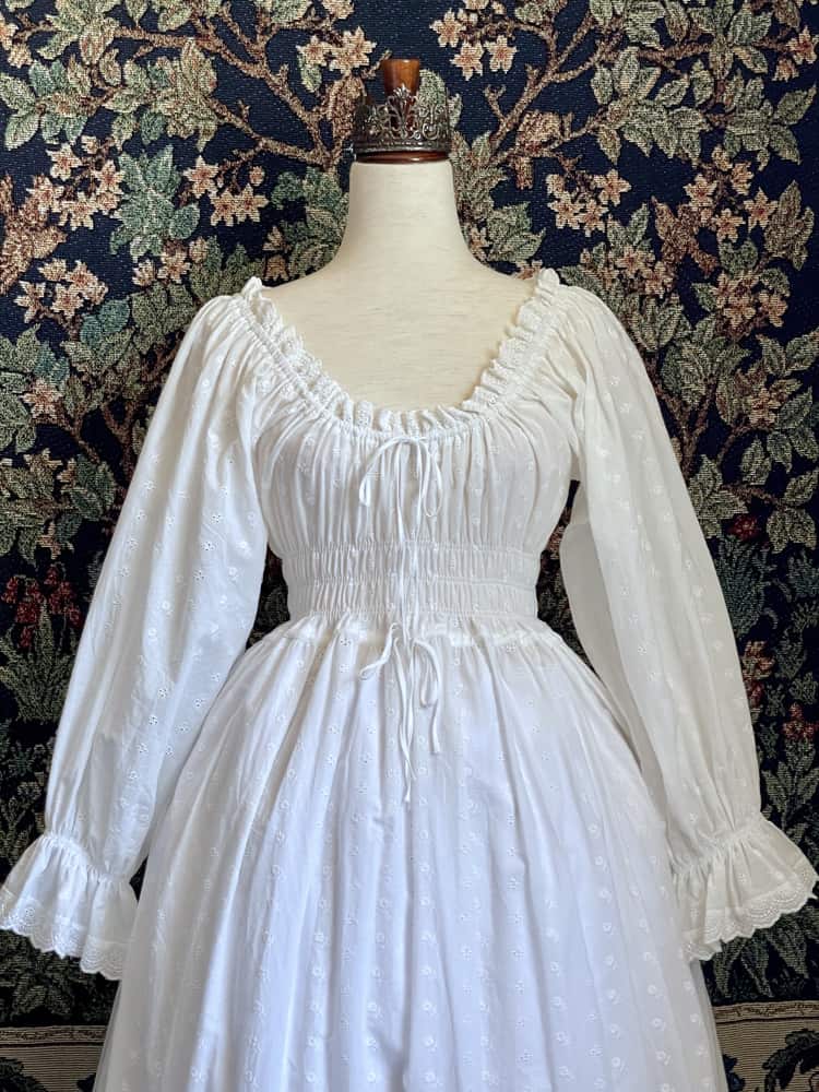 A Historically inspired white 100% cotton smocked chemise dress with bishop bell sleeves is pictured on a mannequin in front of an ornate tapestry background.