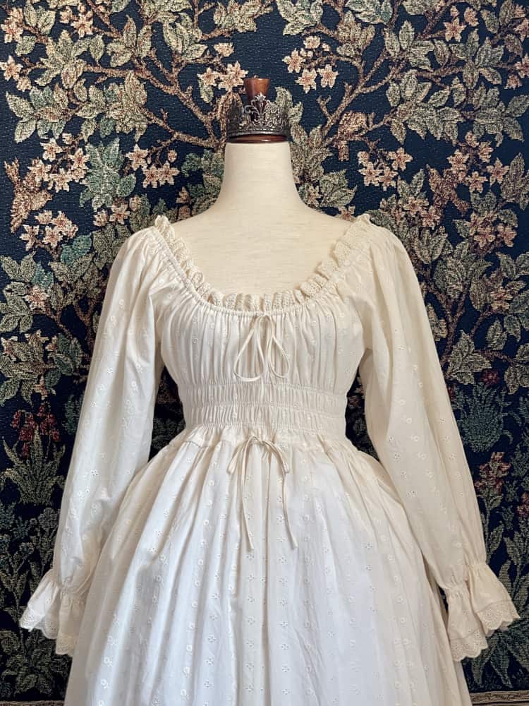 A Historically inspired sheer ivory 100% Cotton smocked chemise maxi dress with bishop bell sleeves is pictured on a mannequin in front of an ornate botanical tapestry.