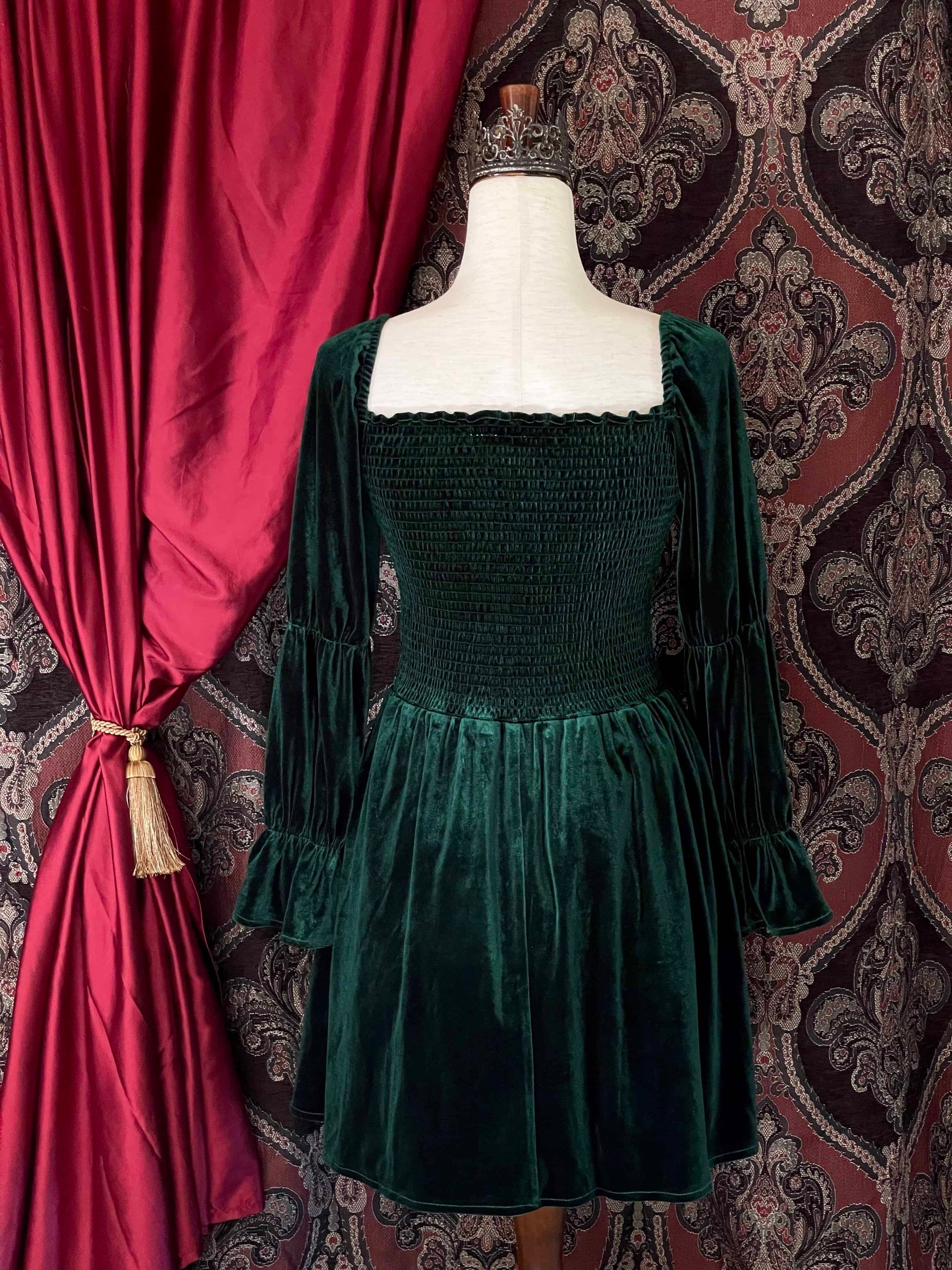 A Renaissance Inspired emerald green velvet smocked mini dress with puff virago sleeves is pictured on a mannequin in front of an ornate background.