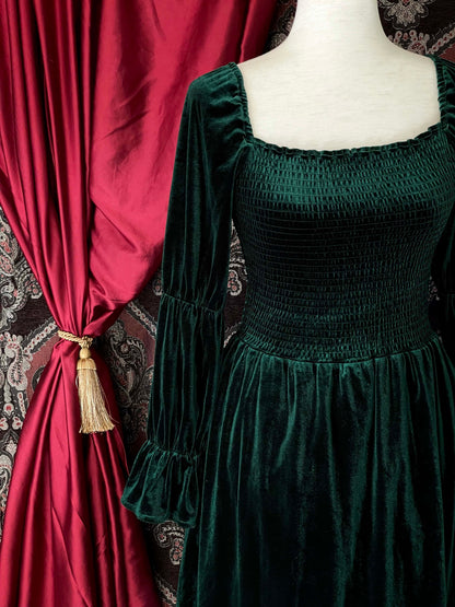 A Renaissance Inspired emerald green velvet smocked mini dress with puff virago sleeves is pictured on a mannequin in front of an ornate background.