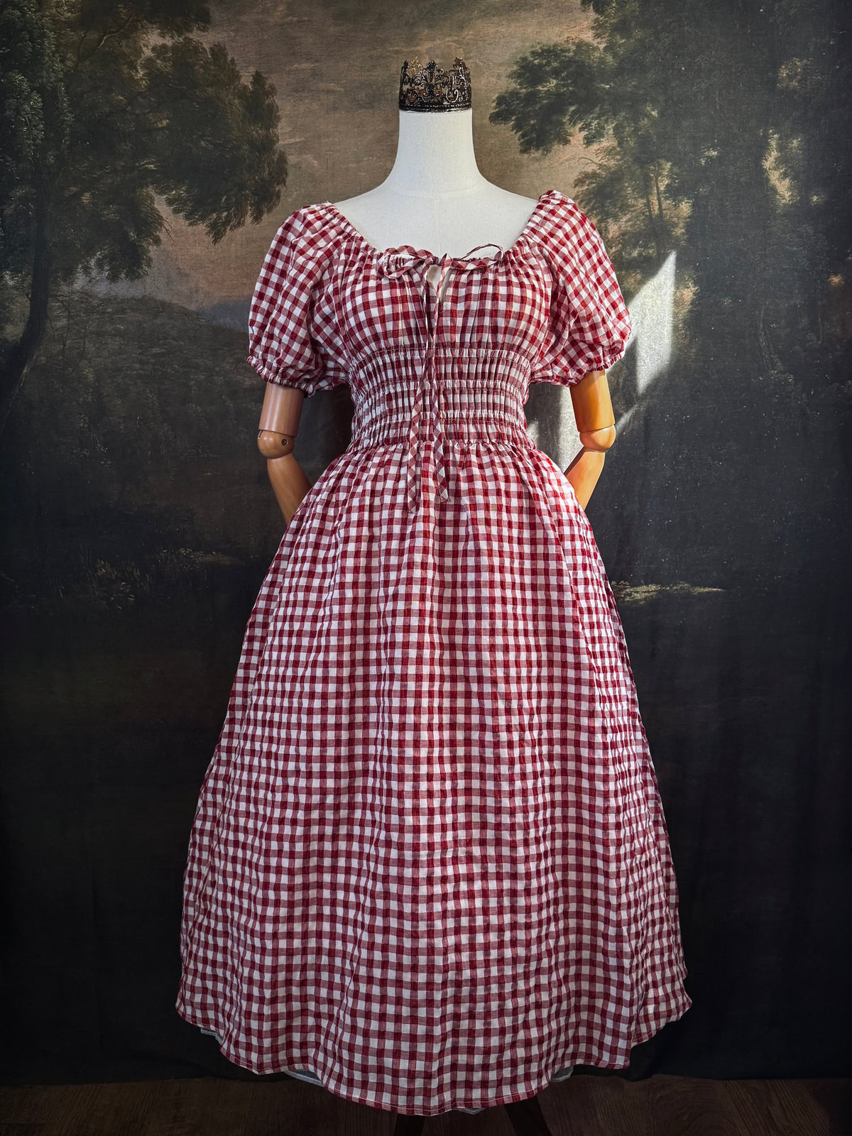 A red and white gingham midi dress with puff sleeves and a smocked waist, displayed on a mannequin, set against a dark vintage-style landscape backdrop.