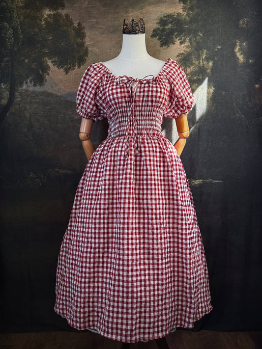A red and white gingham midi dress with puff sleeves and a smocked waist, displayed on a mannequin, set against a dark vintage-style landscape backdrop.