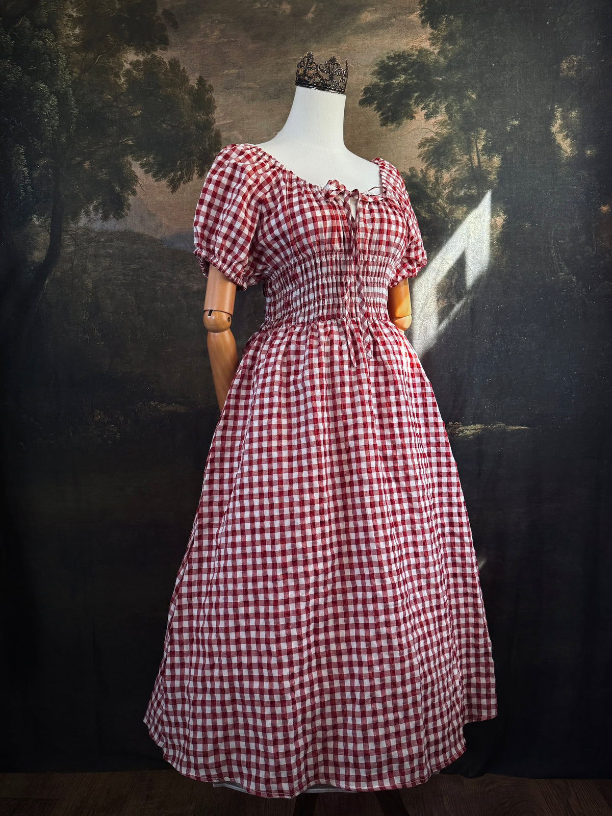 A red and white gingham midi dress with puff sleeves and a smocked waist, displayed on a mannequin, set against a dark vintage-style landscape backdrop.