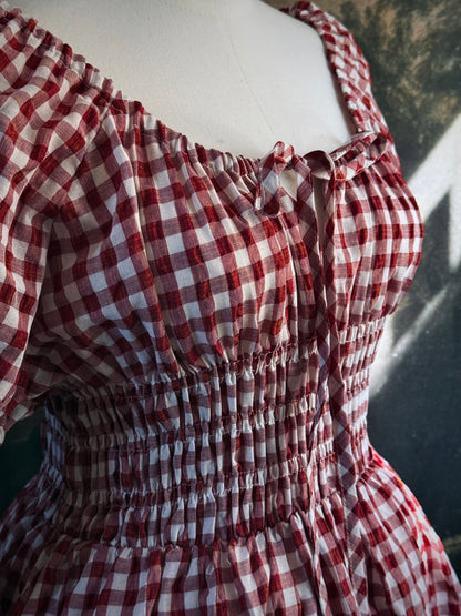 A red and white gingham midi dress with puff sleeves and a smocked waist, displayed on a mannequin, set against a dark vintage-style landscape backdrop.