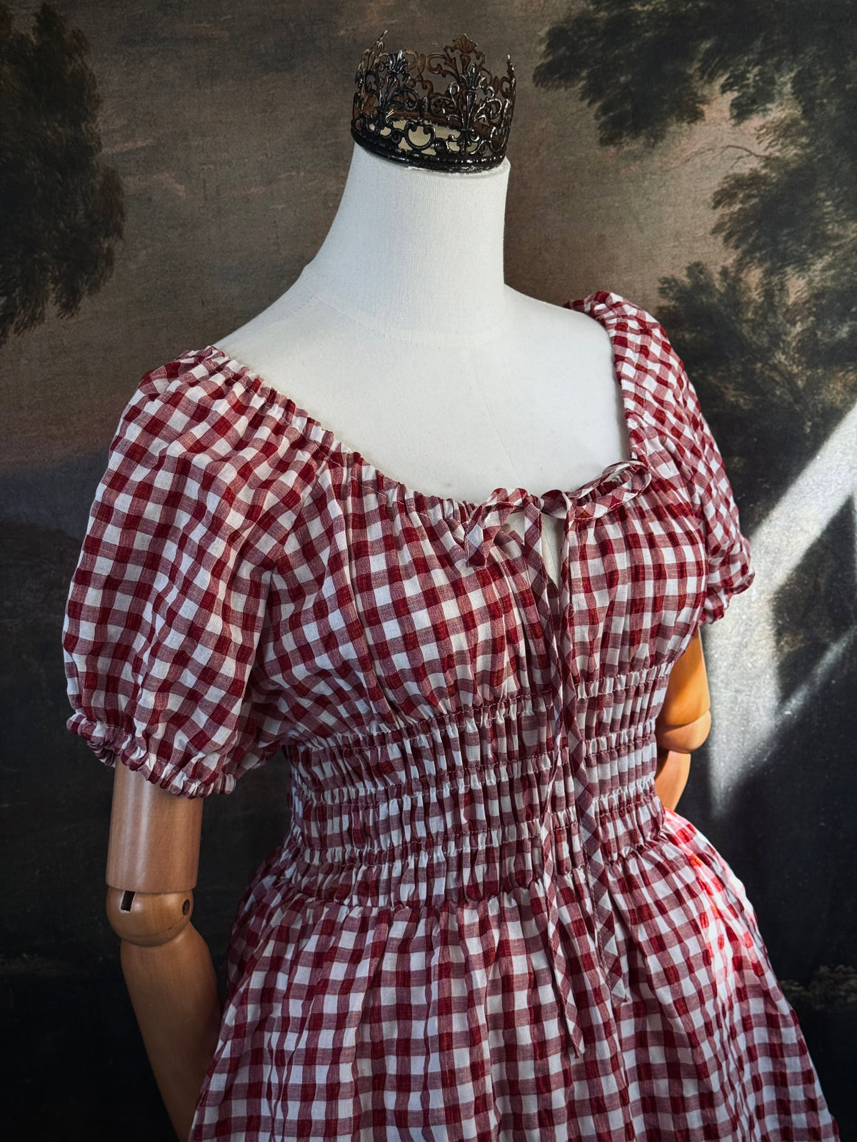 A red and white gingham midi dress with puff sleeves and a smocked waist, displayed on a mannequin, set against a dark vintage-style landscape backdrop.