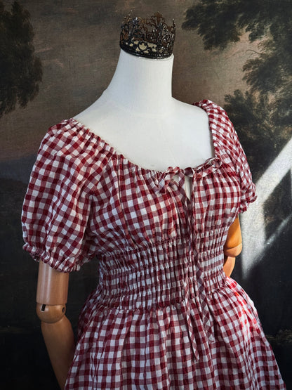 A red and white gingham midi dress with puff sleeves and a smocked waist, displayed on a mannequin, set against a dark vintage-style landscape backdrop.