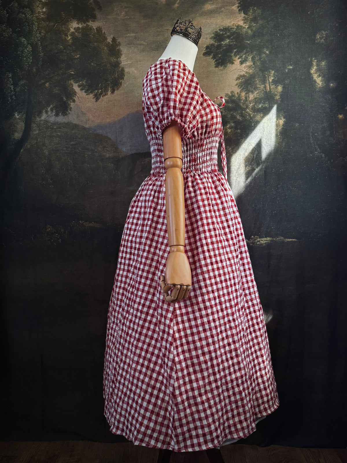 A red and white gingham midi dress with puff sleeves and a smocked waist, displayed on a mannequin, set against a dark vintage-style landscape backdrop.