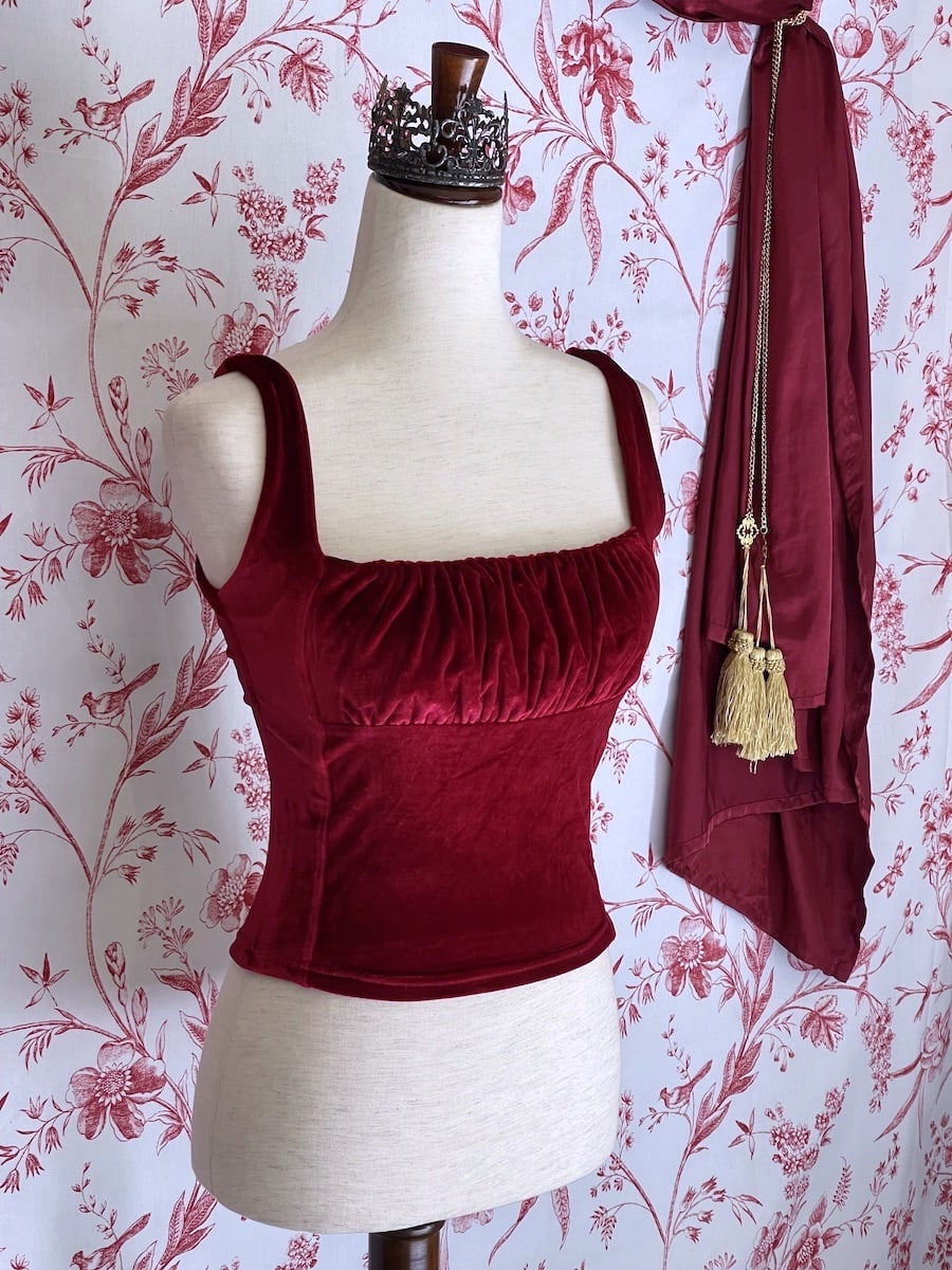 A historically inspired burgundy red velvet ruched milkmaid bustier top is pictured on a mannequin in front of a red and white ornate toile Wallpaper.