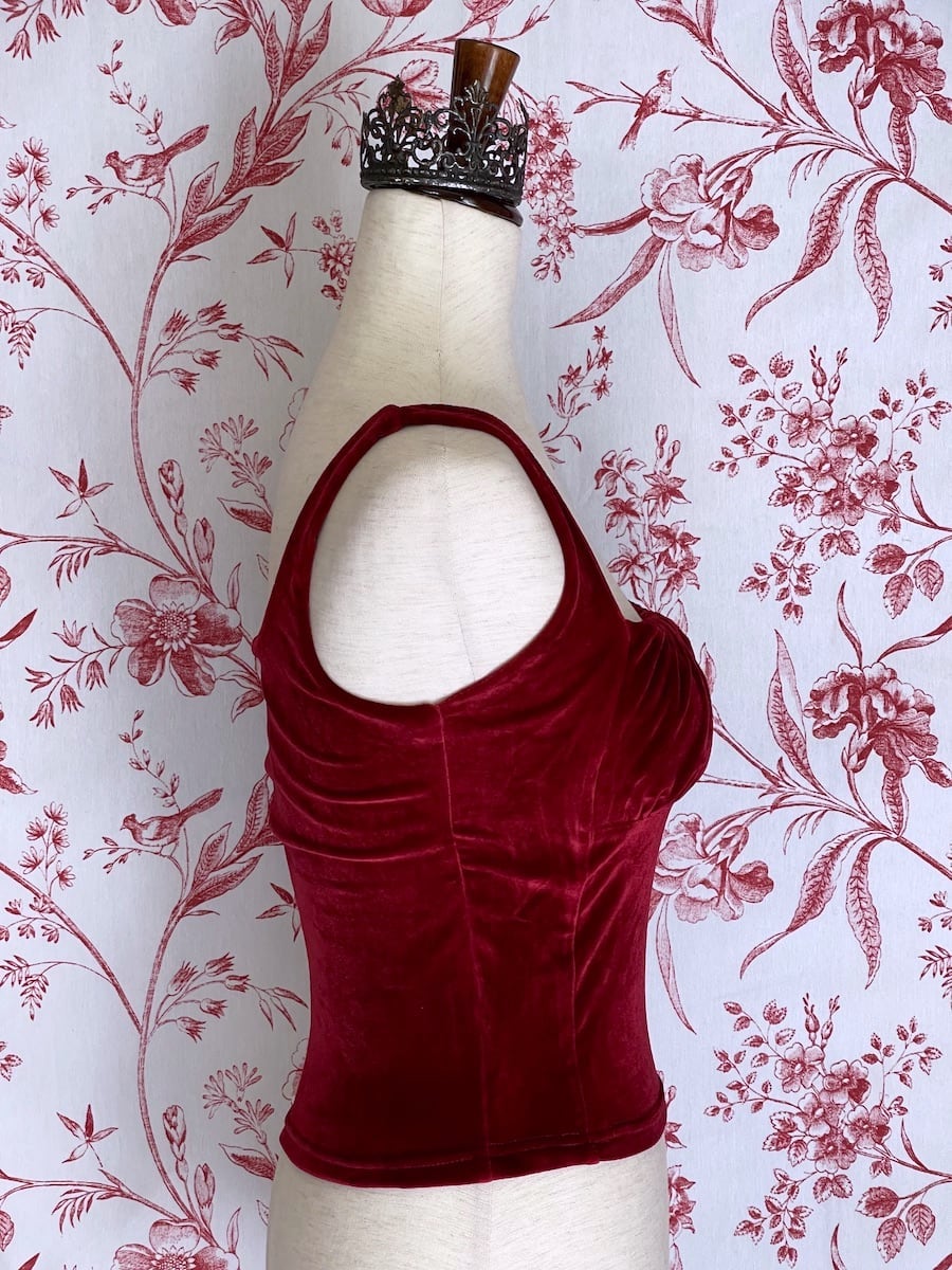 A historically inspired burgundy red velvet ruched milkmaid bustier top is pictured on a mannequin in front of a red and white ornate toile Wallpaper.
