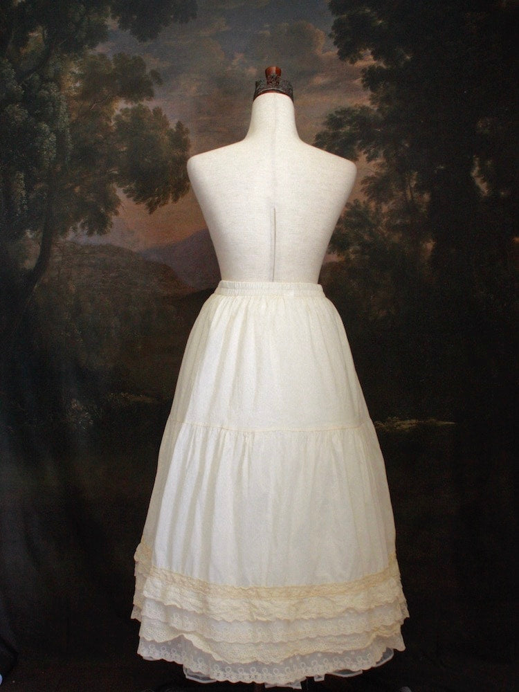 A Historically Inspired bohemian Tiered Cotton Maxi Skirt with floral Lace Trim in Ivory/Cream on a mannequin in front of an art history backdrop.