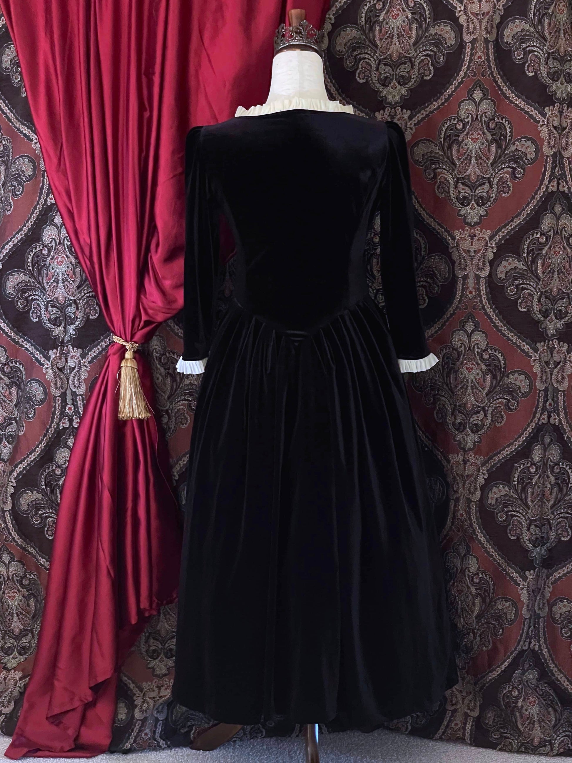 A historically inspired renaissance tudor era gothic black velvet gown with structured corset style bodice and ruffle lace trim, pictured on a mannequin in front of an ornate background.