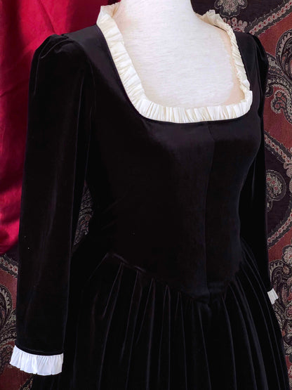 A historically inspired renaissance tudor era gothic black velvet gown with structured corset style bodice and ruffle lace trim, pictured on a mannequin in front of an ornate background.