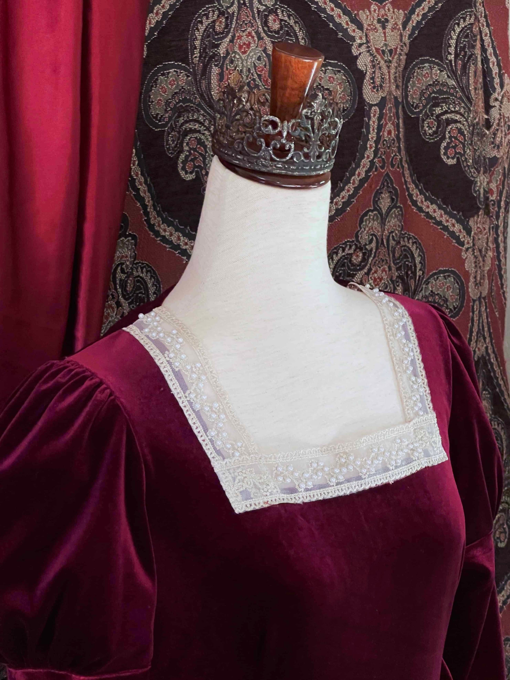 A Renaissance and Tudor inspired Burgundy Velvet Juliet Sleeve Gown with lace trim is pictured on a mannequin in front of a historical ornate backdrop.