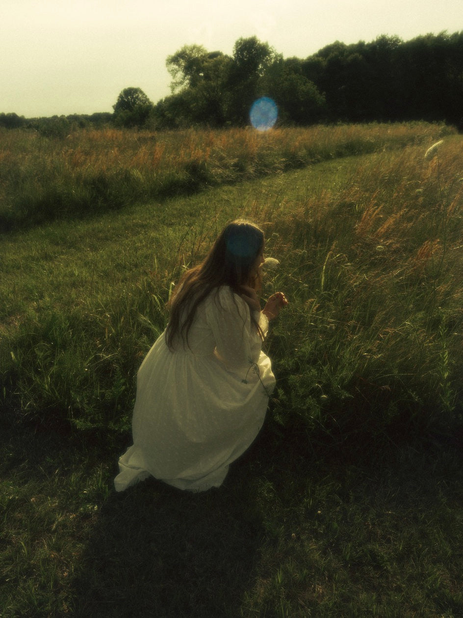 A woman in a field wearing a vintage style historically-inspired white long-sleeve chemise dress with lace trim and gathered details, holding a small white flower cluster.