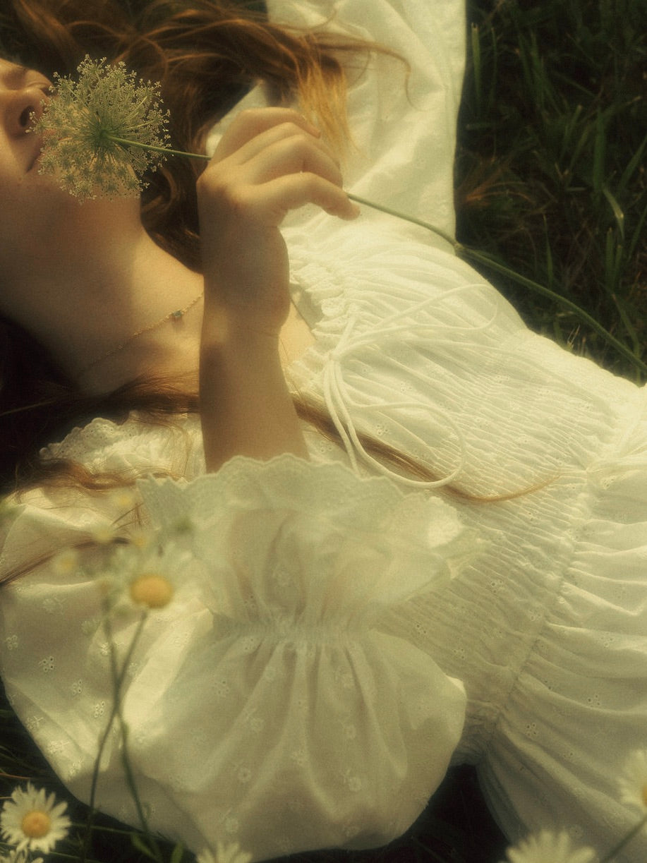 A woman in a field wearing a vintage style historically-inspired white long-sleeve chemise dress with lace trim and gathered details, holding a small white flower cluster.