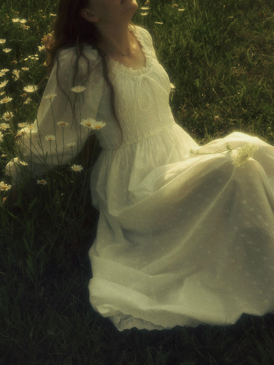 A woman in a long historically-inspired white chemise dress sitting in a grassy field with small white flowers. The dress has a smocked bodice and long sheer sleeves.