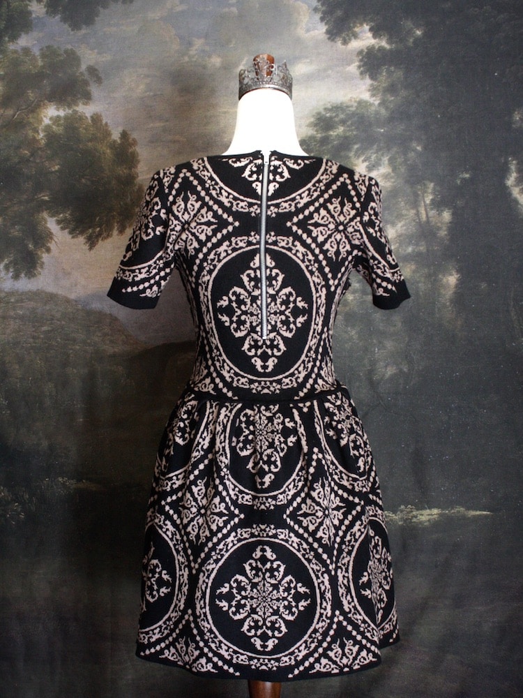A Historically Inspired Ornate Pattern Knit Mini Dress in Black and Gold, pictured on a mannequin in front of an art history backdrop.
