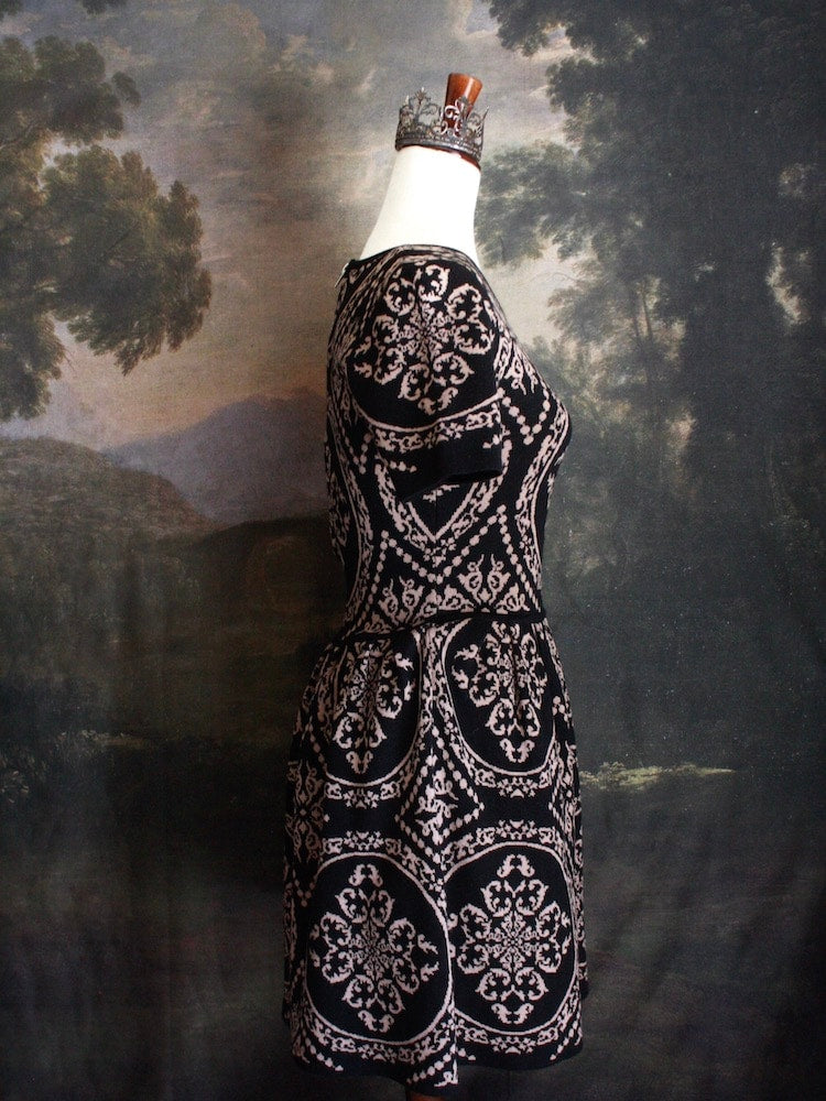 A Historically Inspired Ornate Pattern Knit Mini Dress in Black and Gold, pictured on a mannequin in front of an art history backdrop.
