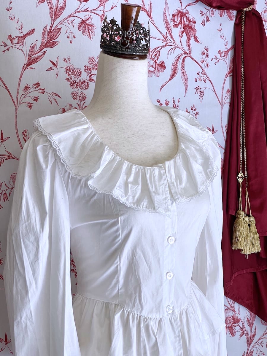 A long sleeved white baroque or victorian ruffle collared blouse inspired by historical fashion is pictured on a mannequin in front of an ornate red and white toile background.