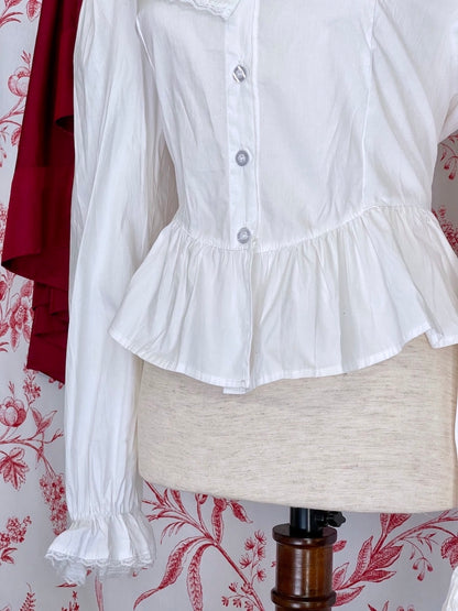 A long sleeved white baroque or victorian ruffle collared blouse inspired by historical fashion is pictured on a mannequin in front of an ornate red and white toile background.