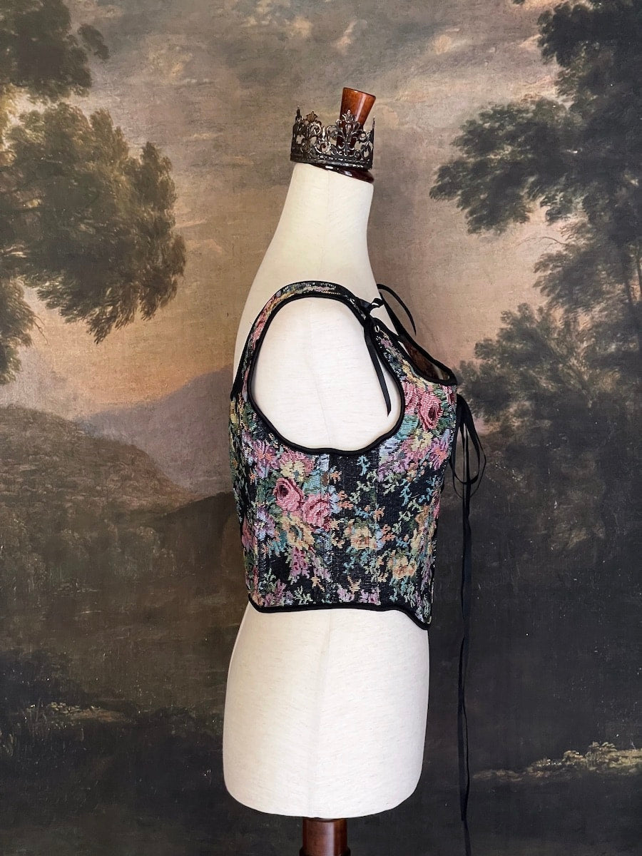 A historically inspired black floral print woven gobelin tapestry corset is pictured on a mannequin in front of an ornate  landscape painting wallpaper.