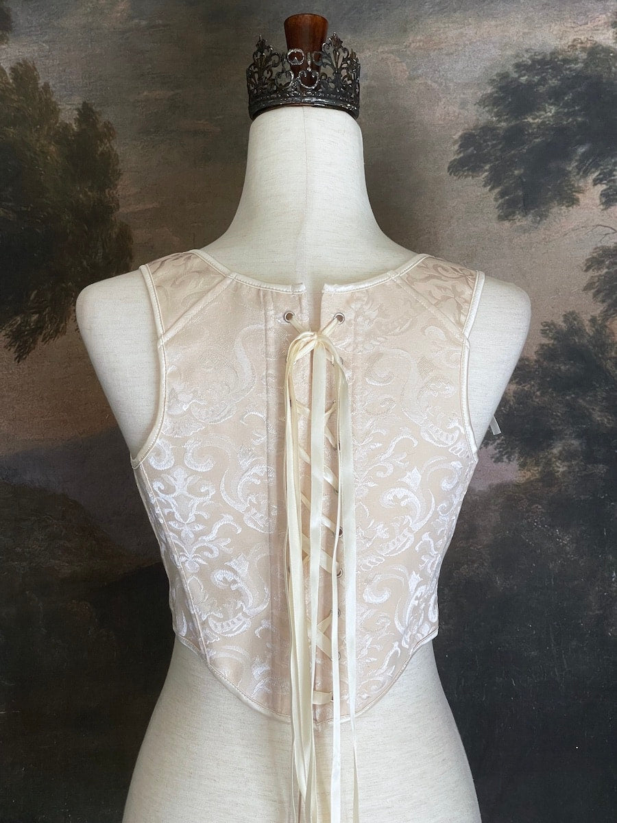 A Historically inspired Tudor, baroque, or renaissance era corset with champagne-ivory colored ornate jacquard fabric is pictured on a mannequin in front of a historic painted landscape backdrop.