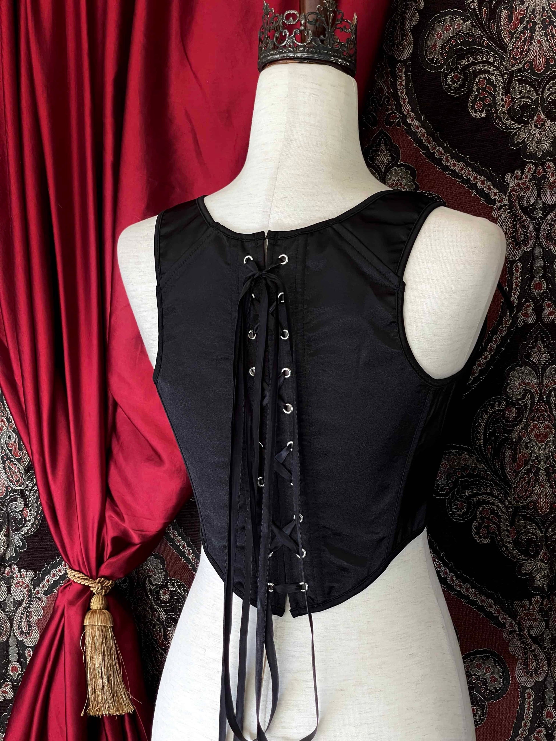 A Gothic Black Satin Renaissance Tudor Era Corset or Pair of stays is pictured on a mannequin in front of a historical Ornate Backdrop.