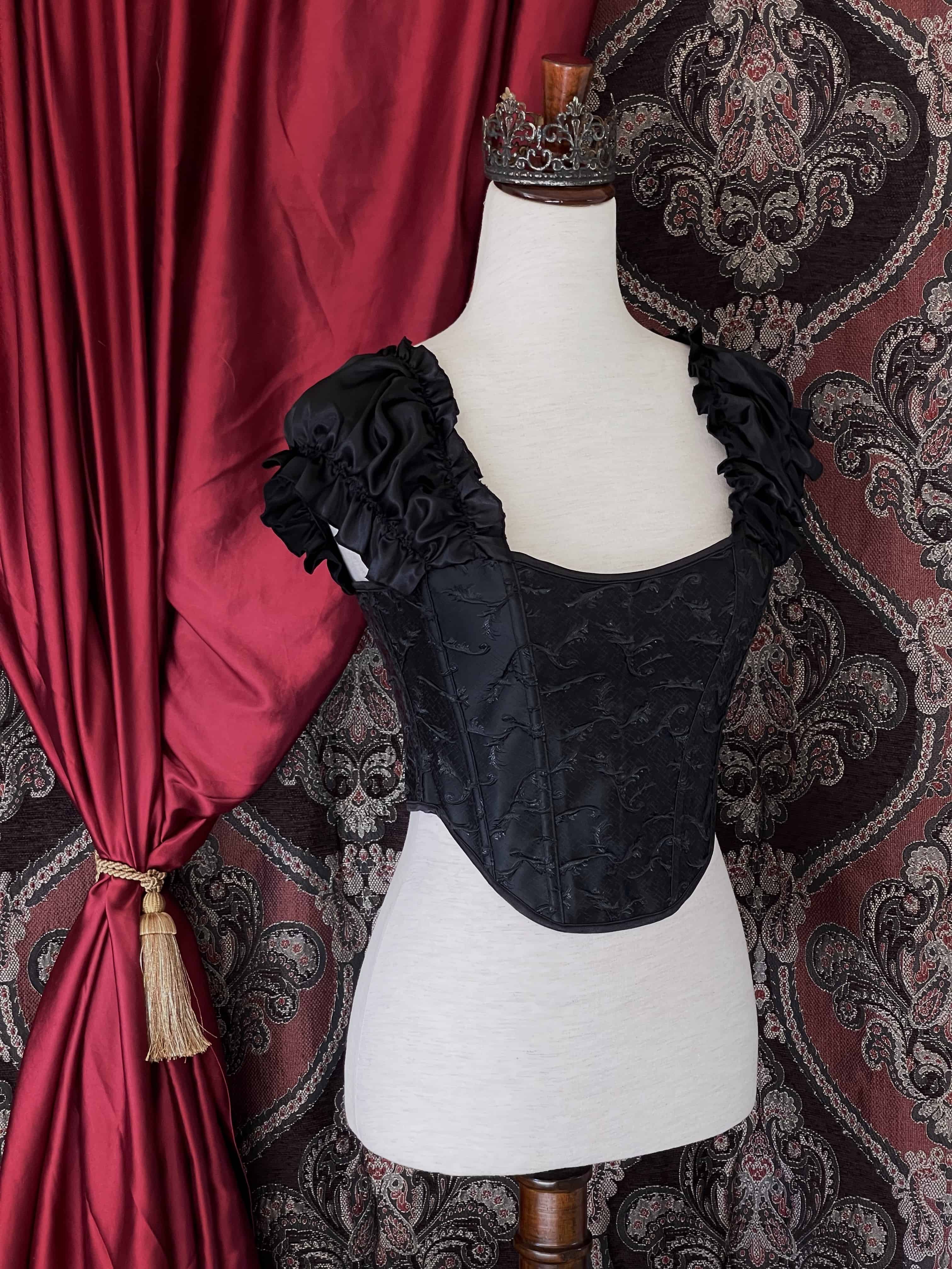 A historically inspired renaissance and baroque era corset  made of black embroidered fabric and puff sleeves is pictured on a mannequin in front of an ornate backdrop.