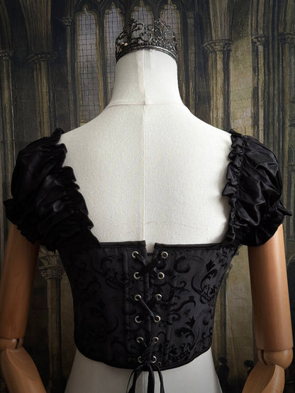 A historically inspired renaissance and baroque era corset made of gothic black jacquard fabric with ruched puff sleeves is pictured on a mannequin in front of a historic cathedral backdrop.