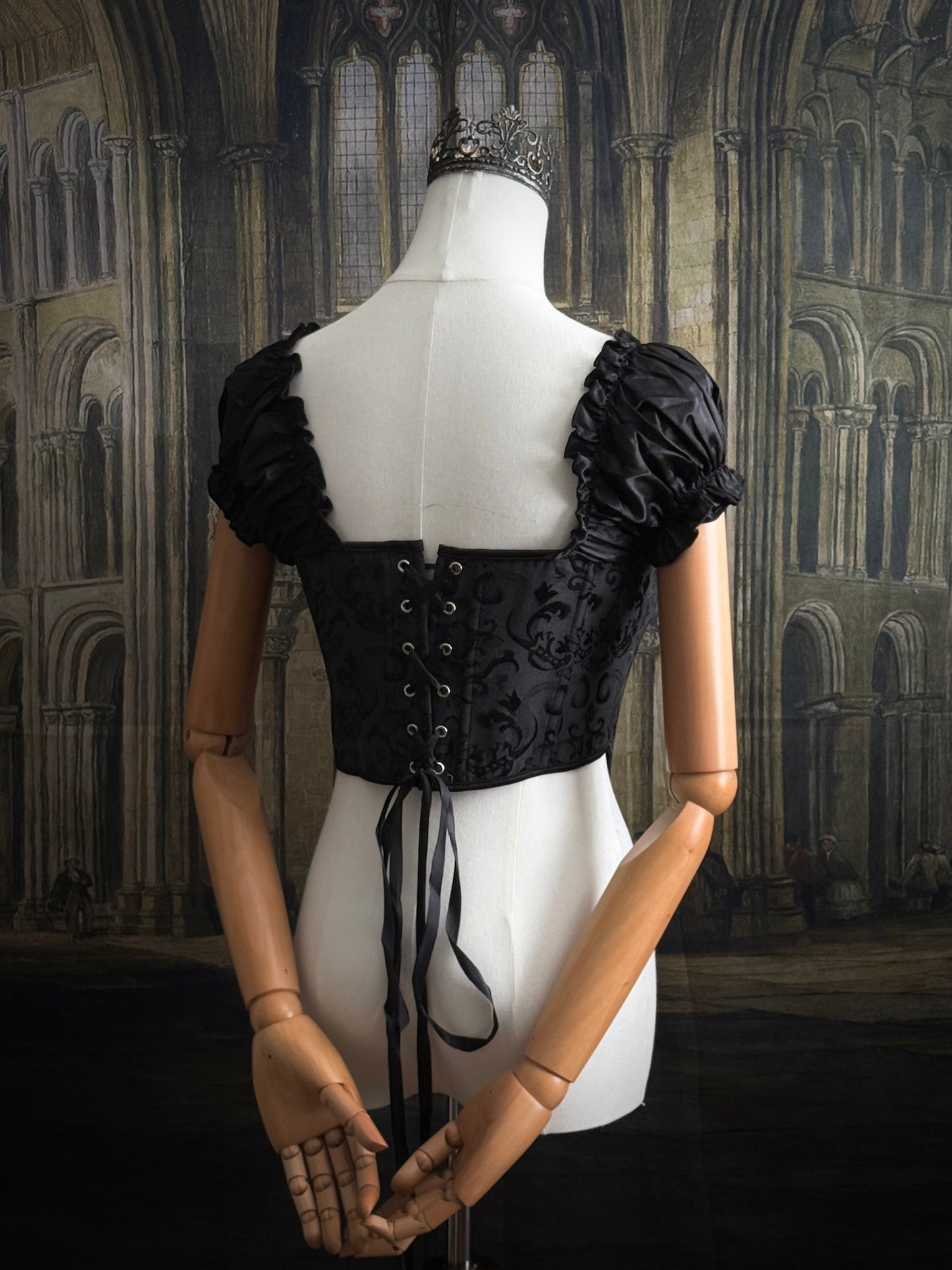 A historically inspired renaissance and baroque era corset made of gothic black jacquard fabric with ruched puff sleeves is pictured on a mannequin in front of a historic cathedral backdrop.