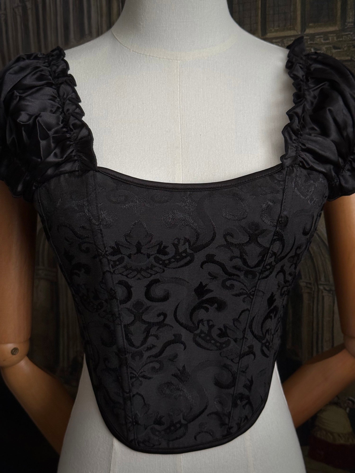 A historically inspired renaissance and baroque era corset made of gothic black jacquard fabric with ruched puff sleeves is pictured on a mannequin in front of a historic cathedral backdrop.