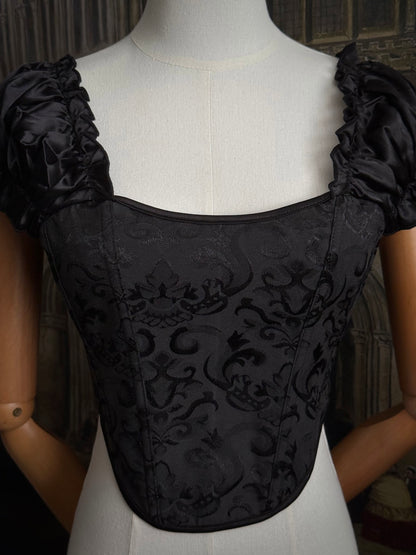 A historically inspired renaissance and baroque era corset made of gothic black jacquard fabric with ruched puff sleeves is pictured on a mannequin in front of a historic cathedral backdrop.