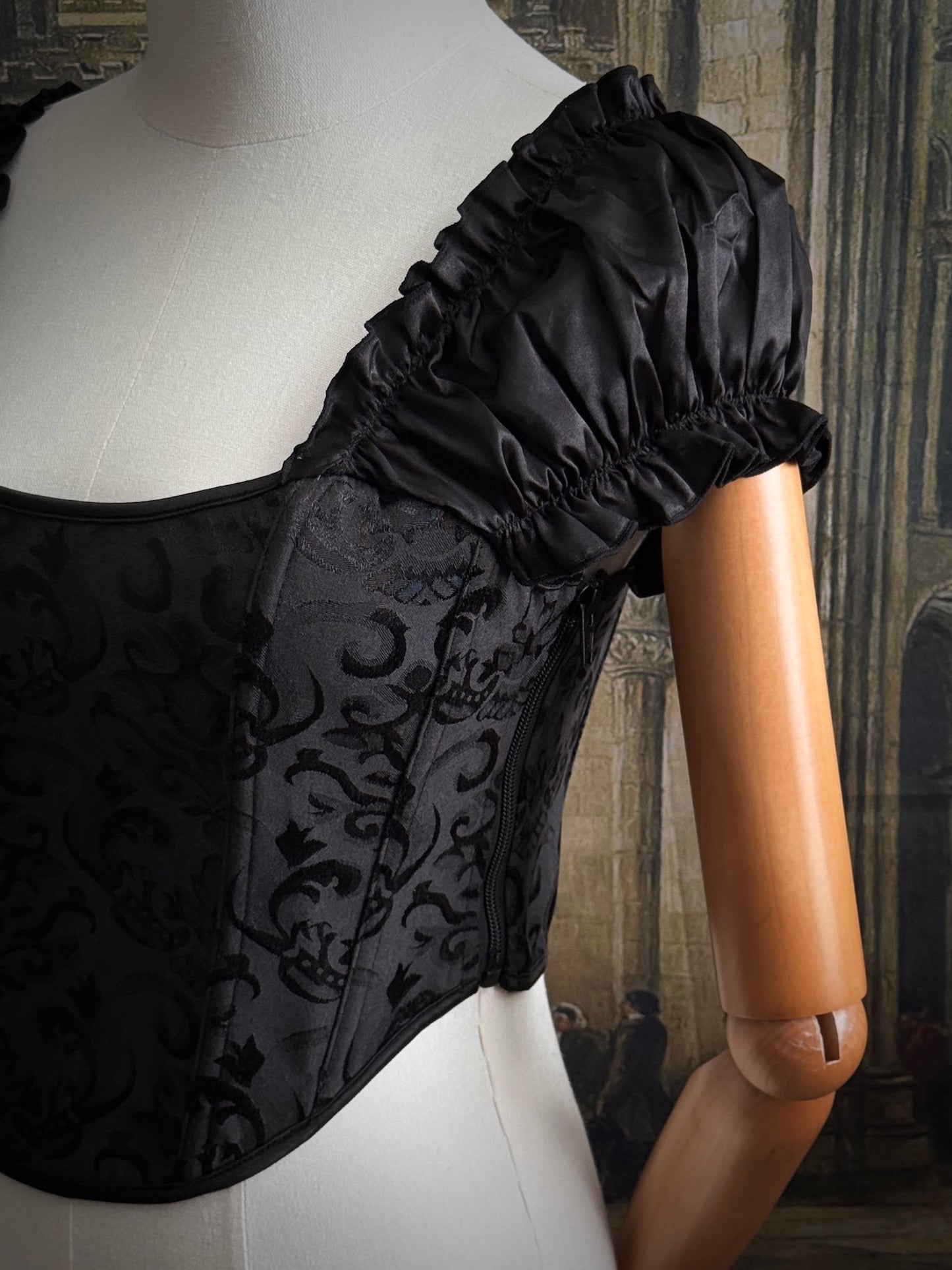 Details of a historically inspired renaissance and baroque era corset made of gothic black jacquard fabric with ruched puff sleeves is pictured on a mannequin in front of a historic cathedral backdrop.