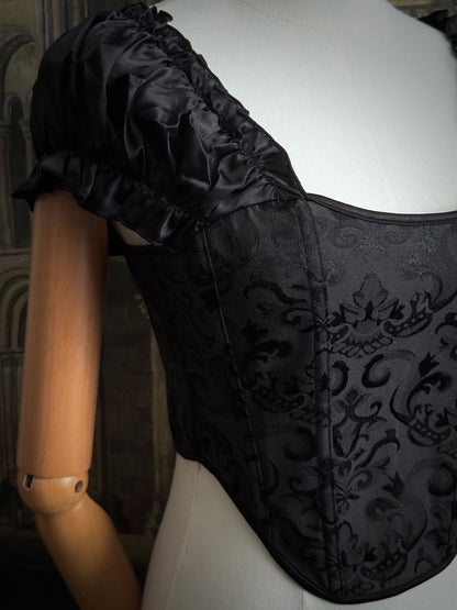 Details of a historically inspired renaissance and baroque era corset made of gothic black jacquard fabric with ruched puff sleeves is pictured on a mannequin in front of a historic cathedral backdrop.
