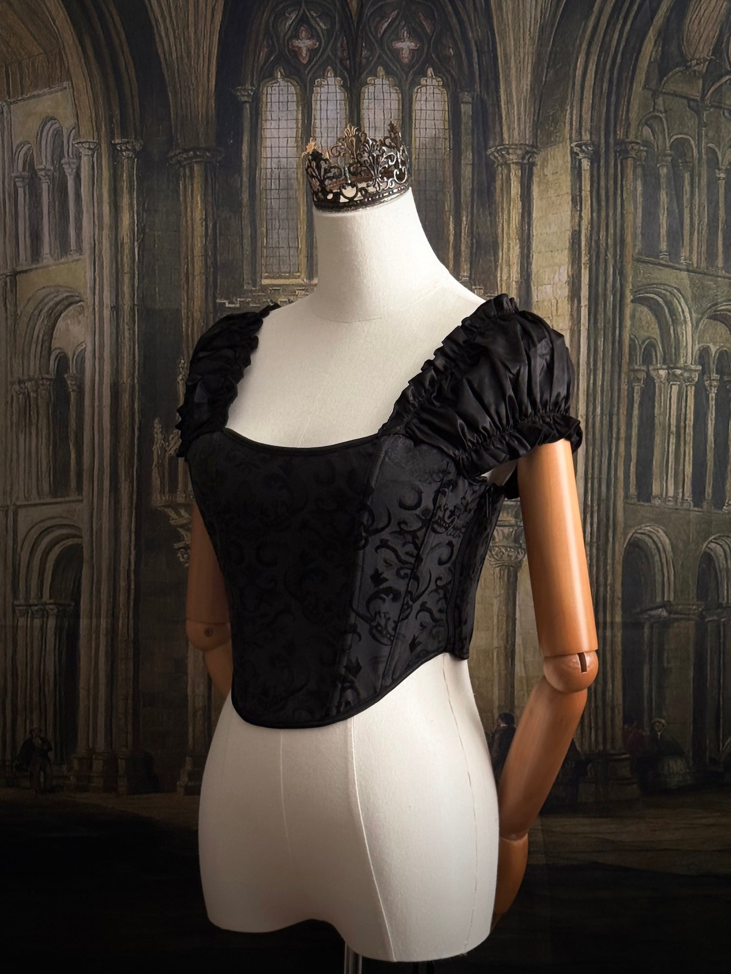 A historically inspired renaissance and baroque era corset made of gothic black jacquard fabric with ruched puff sleeves is pictured on a mannequin in front of a historic cathedral backdrop.