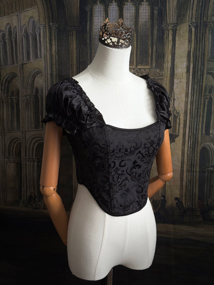 A historically inspired renaissance and baroque era corset made of gothic black jacquard fabric with ruched puff sleeves is pictured on a mannequin in front of a historic cathedral backdrop.