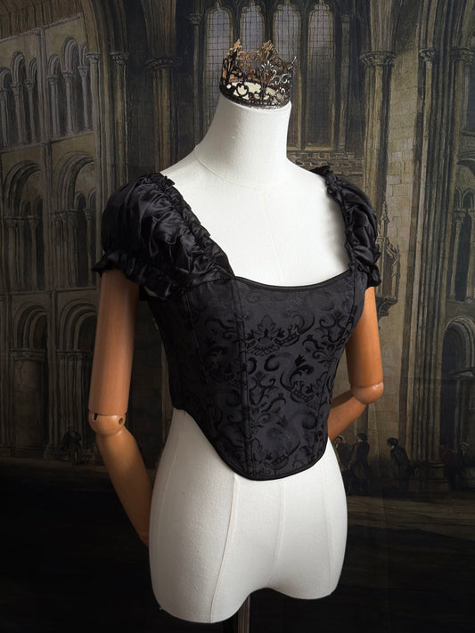 A historically inspired renaissance and baroque era corset made of gothic black jacquard fabric with ruched puff sleeves is pictured on a mannequin in front of a historic cathedral backdrop.