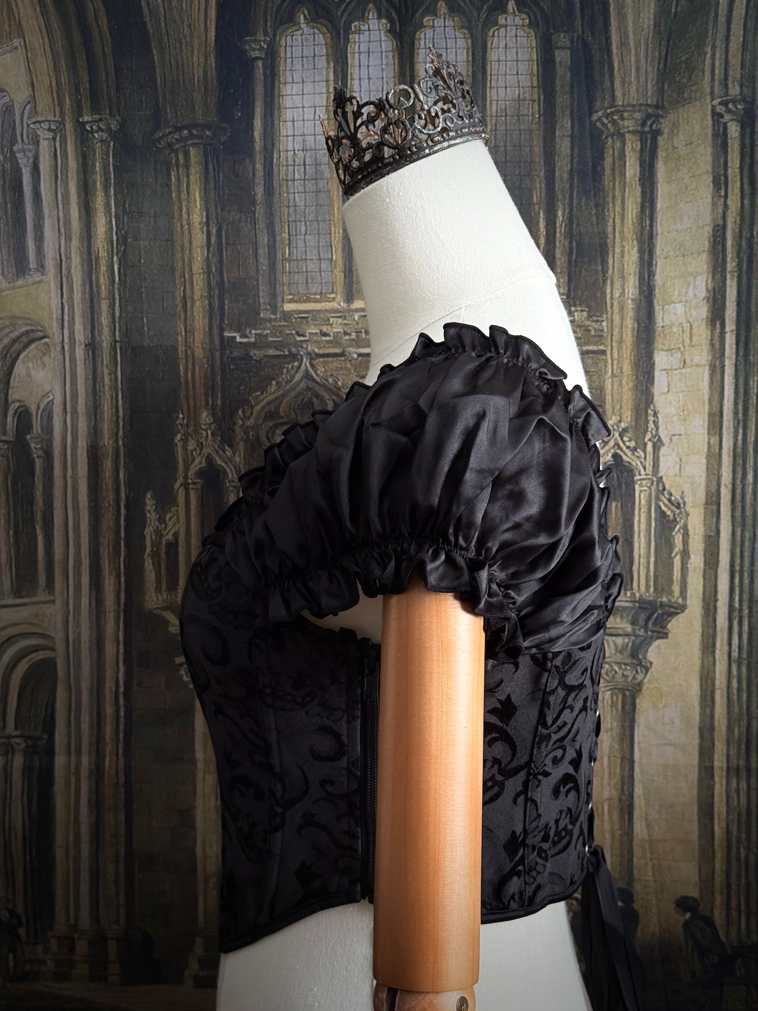 A historically inspired renaissance and baroque era corset made of gothic black jacquard fabric with ruched puff sleeves is pictured on a mannequin in front of a historic cathedral backdrop.