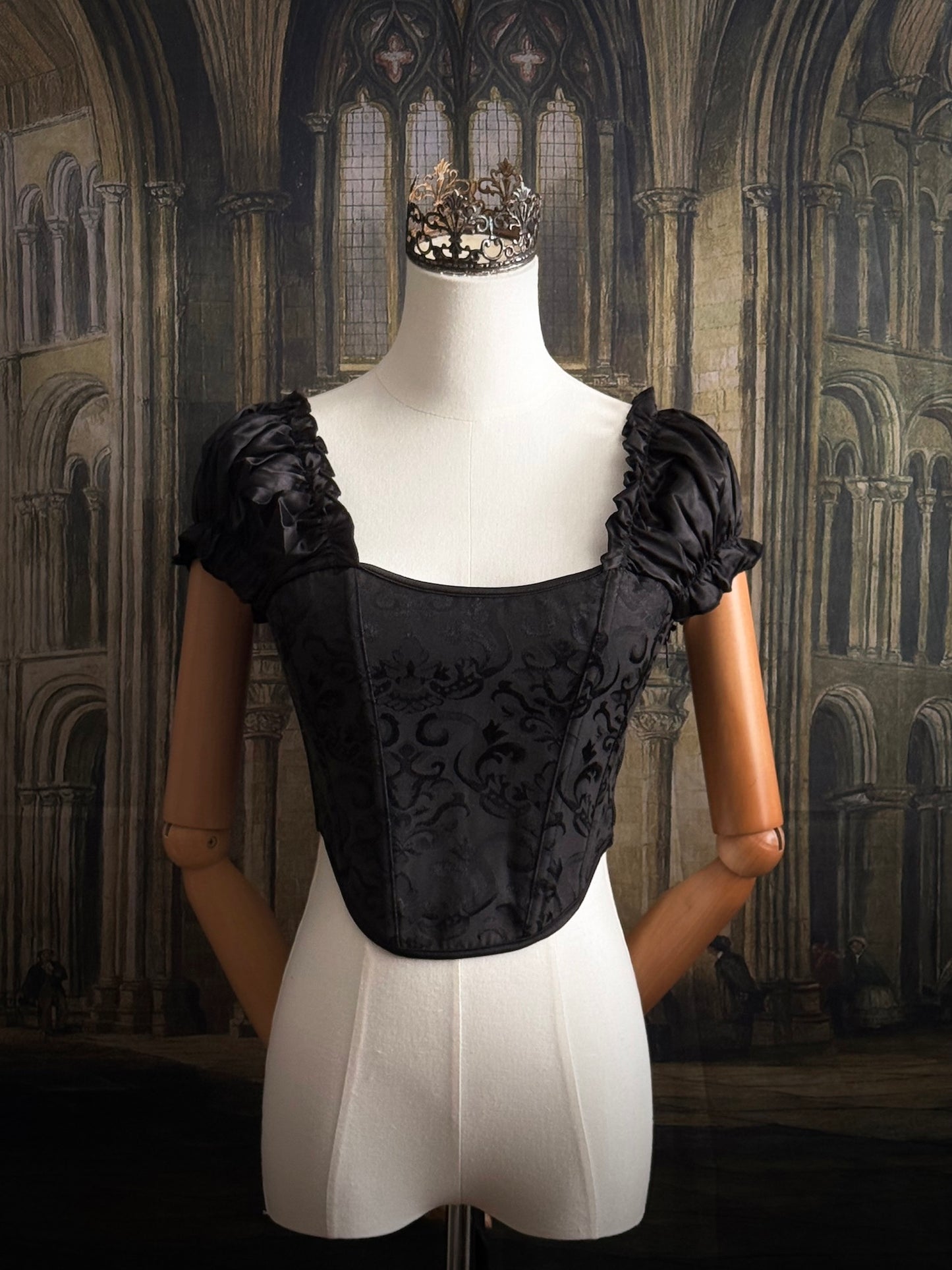 A historically inspired renaissance and baroque era corset made of gothic black jacquard fabric with ruched puff sleeves is pictured on a mannequin in front of a historic cathedral backdrop.
