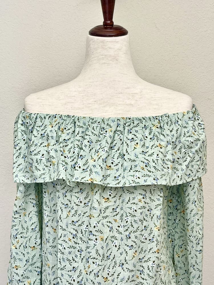 A light mint green floral print off the shoulder blouse inspired by victorian era fashion is pictured on a mannequin.