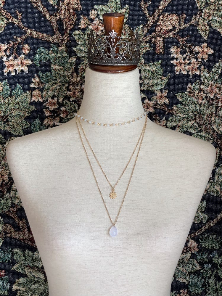 A gold colored stacked fantasy necklace with crystal pendant and sun charm is pictured on a mannequin.