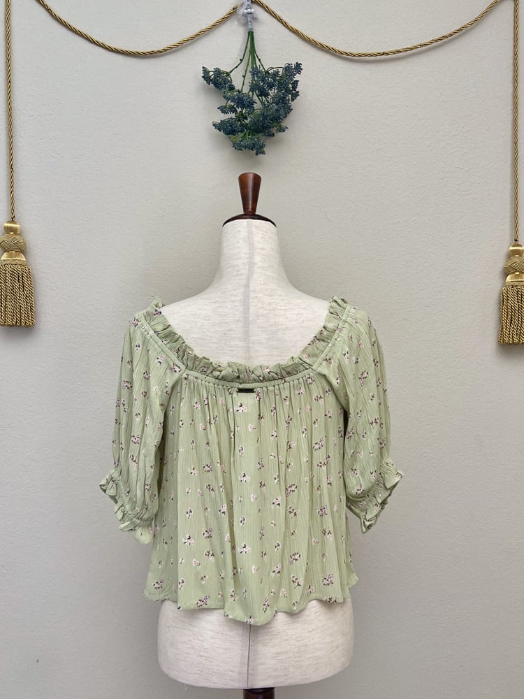 A Sage Green Historically Inspired Short Sleeve Peasant Blouse with Tie Front Ruched Neckline is pictured on a mannequin in front of a classical background.
