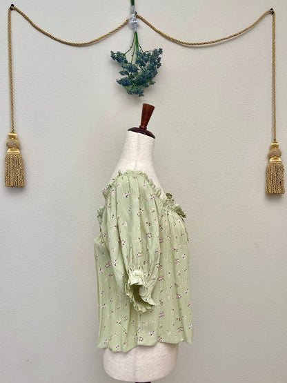 A Sage Green Historically Inspired Short Sleeve Peasant Blouse with Tie Front Ruched Neckline is pictured on a mannequin in front of a classical background.
