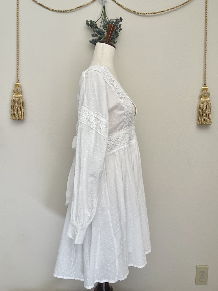 A Victorian or Edwardian inspired white cotton vneck bishop sleeve open back dress with crocheted lace details is pictured on a mannequin in front of a historically inspired background.