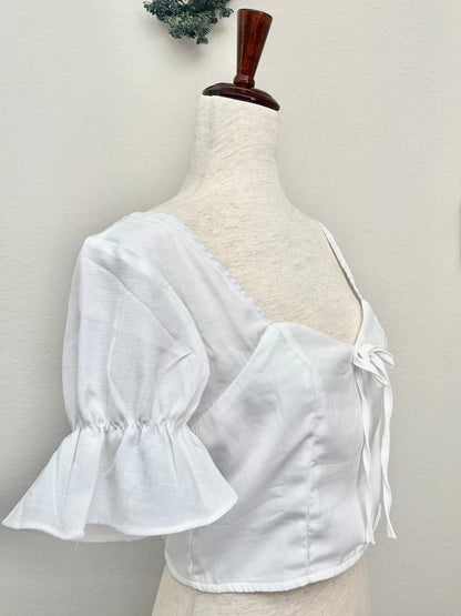 A white edwardian era inspired cropped blouse with ruffle sleeves is pictured on a mannequin in front of a classical background.